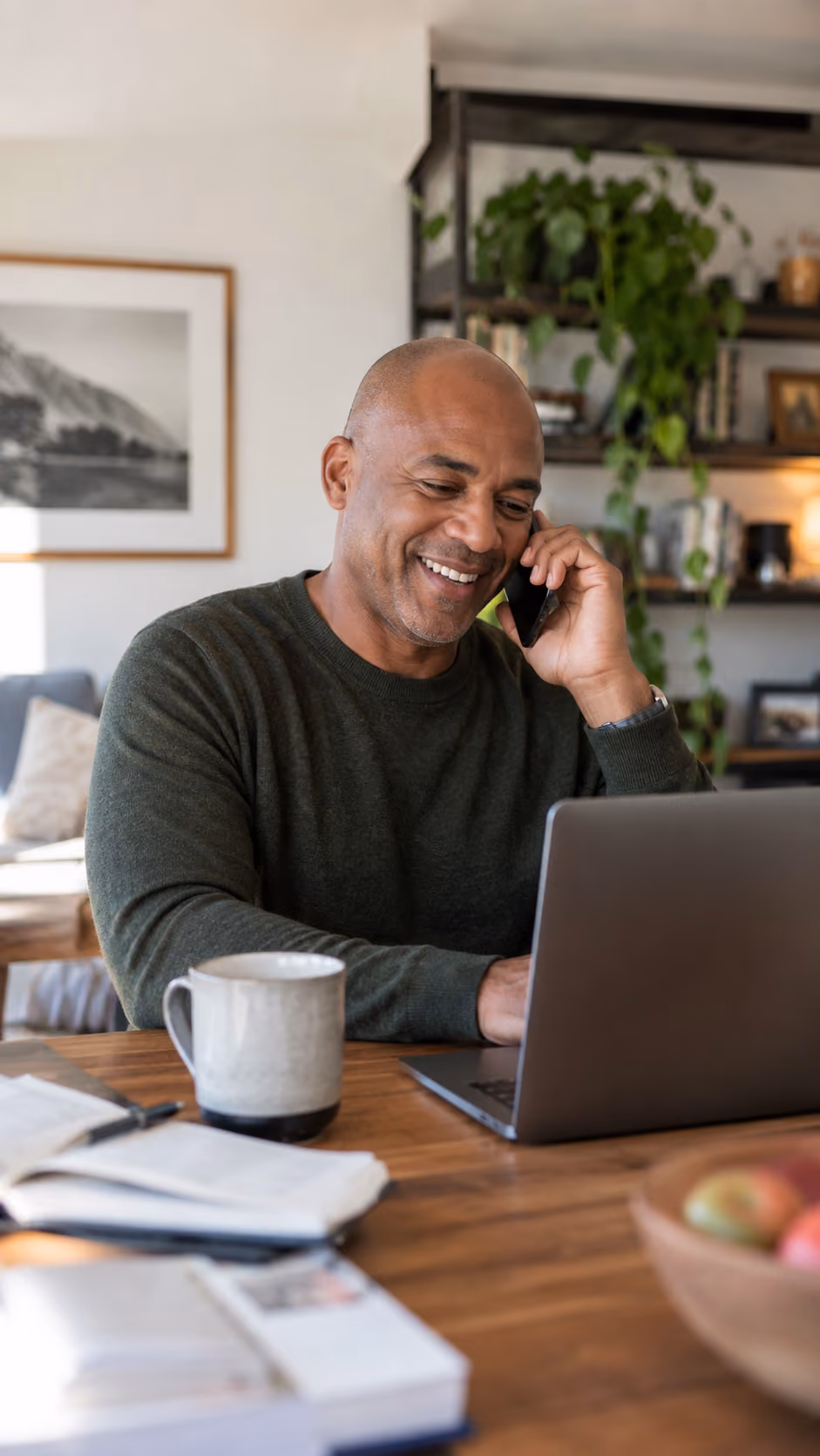 Smiling man in a dark green sweater sitting at a wooden table, talking on a phone while using a laptop with a coffee mug and books nearby in a cozy home with plants and framed pictures in the background.