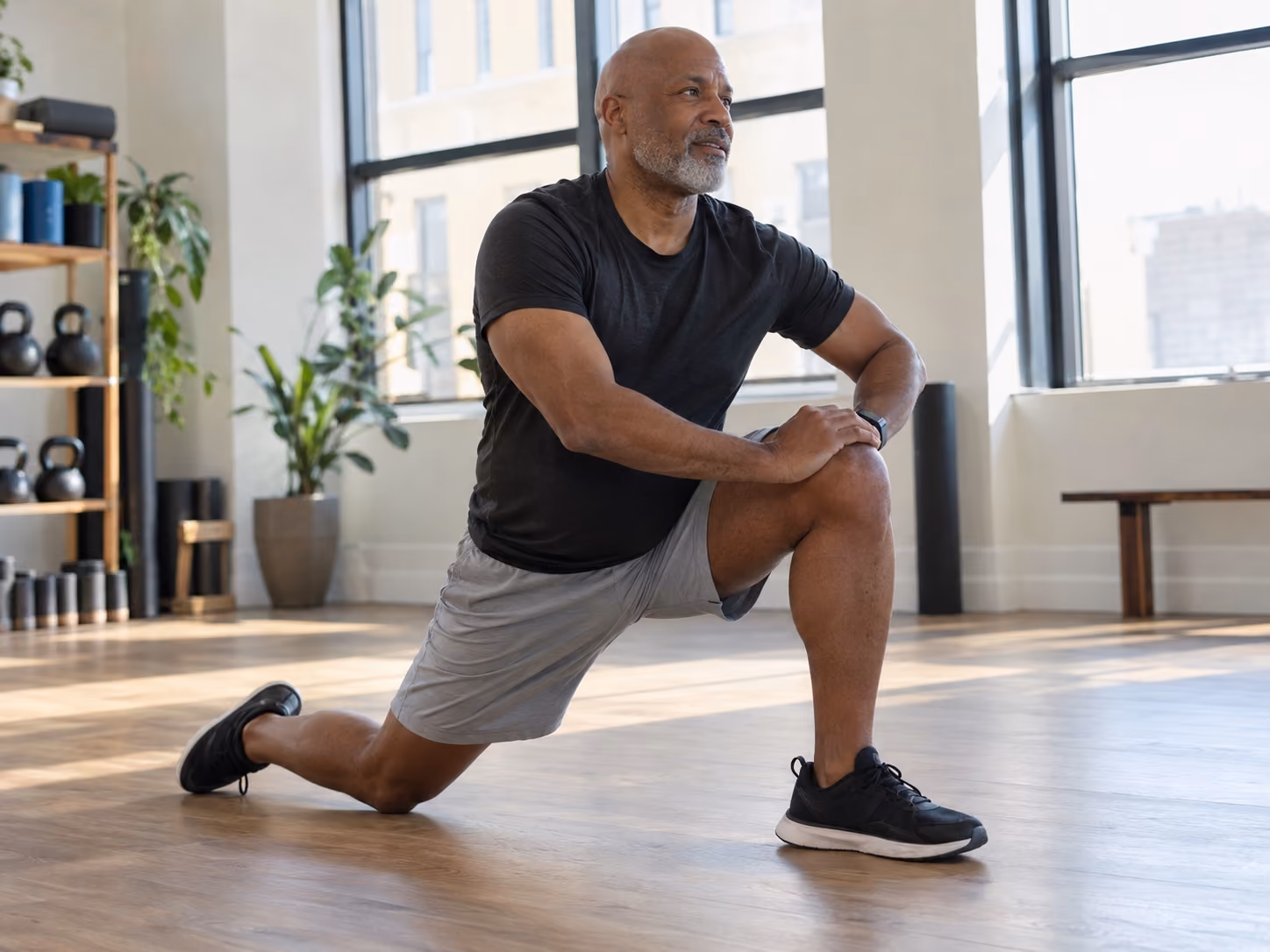 Man in black t-shirt and gray shorts stretching one leg forward on a wooden floor in a bright room with large windows and plants.