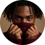 Person with dreadlocks covering lower face with maroon sweater, looking intently at the camera.