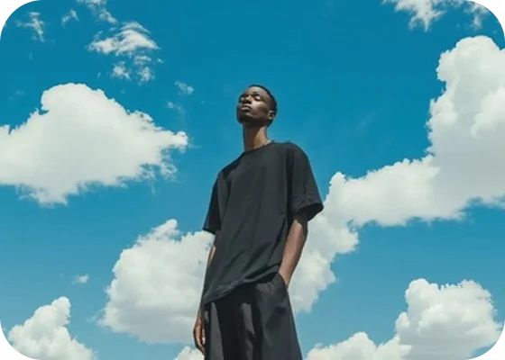 A young man in a black oversized t-shirt and shorts stands outdoors against a bright blue sky with scattered white clouds, looking upwards with a calm expression.