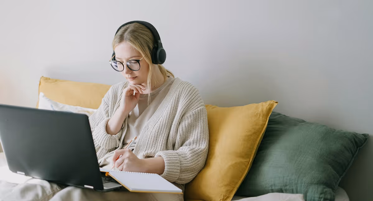 Woman wearing headphones and glasses, working on a laptop while writing in a notebook, seated on a bed with yellow and green pillows.