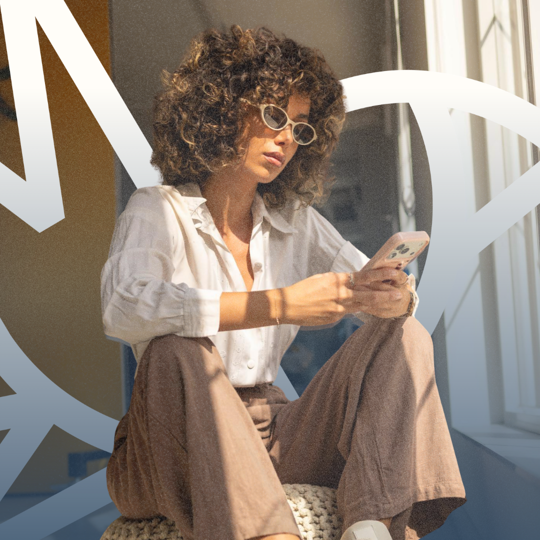 A woman sitting on an ottoman looking at a smartphone.