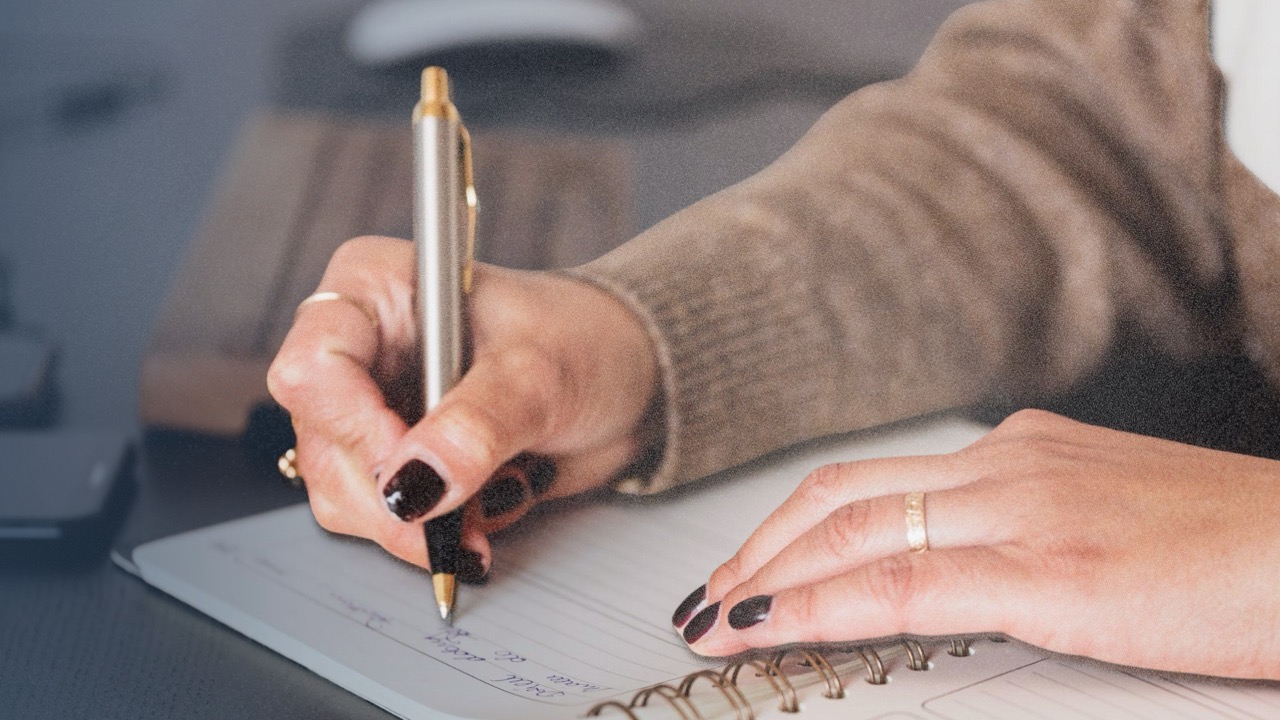 Hands writing a plan in a spiral notebook at a desk.