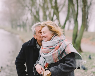 Smiling elderly couple standing close together outdoors with greenery in background.