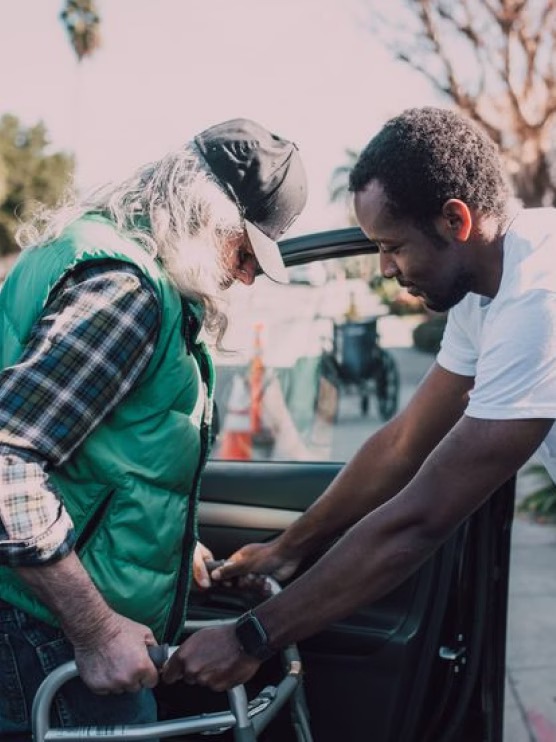 Younger man helping elderly man with walker get into car on sunny day outdoors.