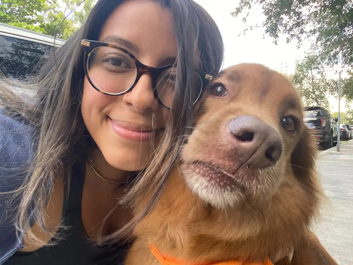 A smiling person wearing glasses beside a brown dog outdoors