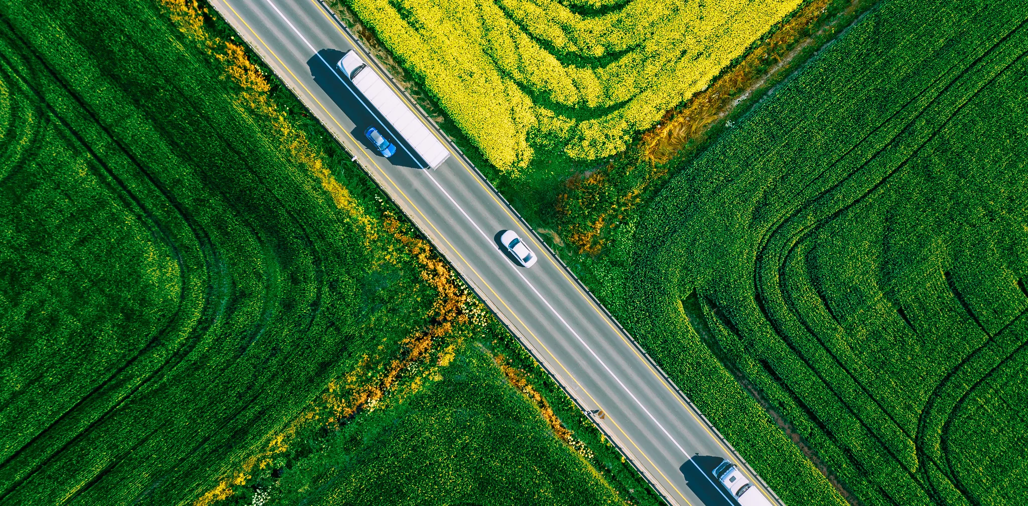 Aerial view of a two-lane road crossing green and yellow fields, with a truck and two white cars
