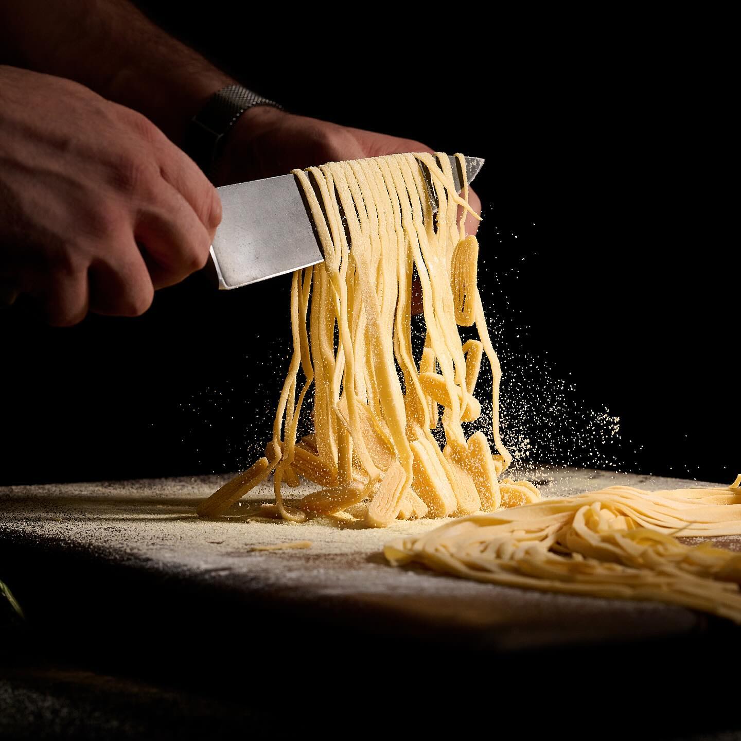 Hands hand-cutting fresh pasta noodles with a knife on a floured board.