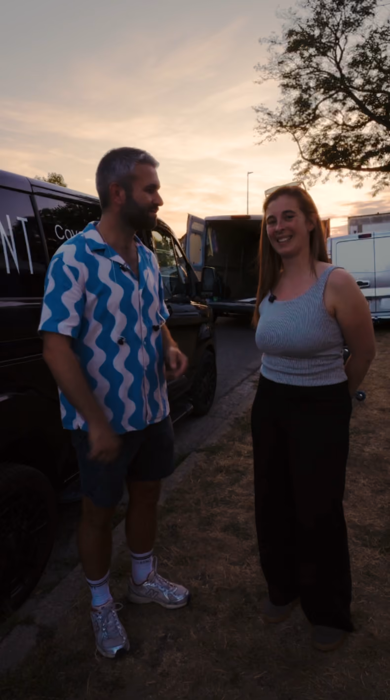 Twee mensen, een man in een blauw-wit golvend patroon overhemd en korte broek en een vrouw in een grijze top en zwarte broek, staan buiten bij een geparkeerd voertuig tijdens zonsondergang.