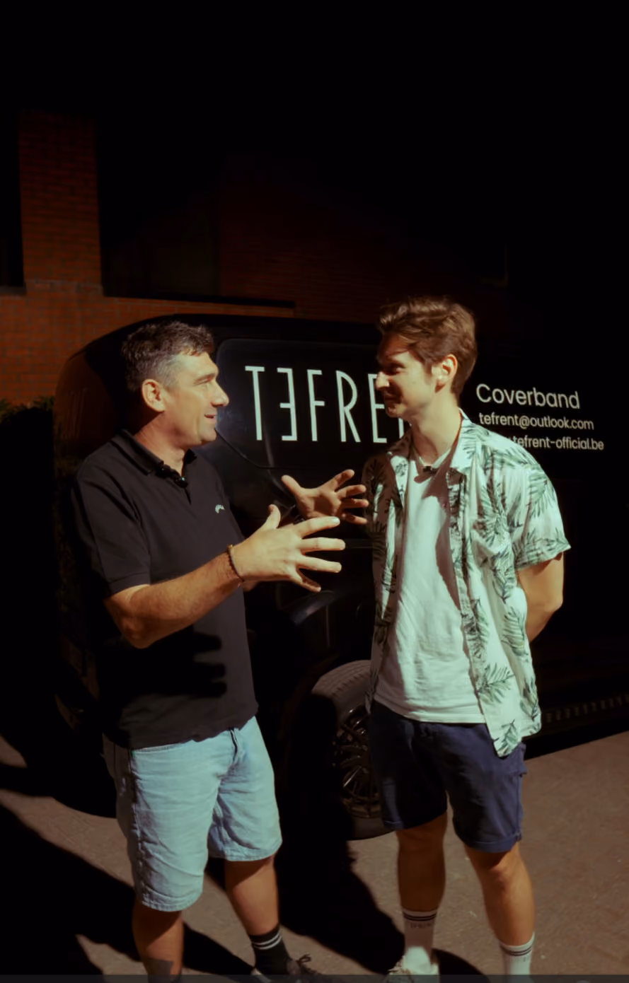 Twee mannen die 's nachts voor een zwarte bestelwagen met het logo van coverband TEFRE en contactinformatie praten, waarbij de man links met zijn handen spreekt en de man rechts luistert.