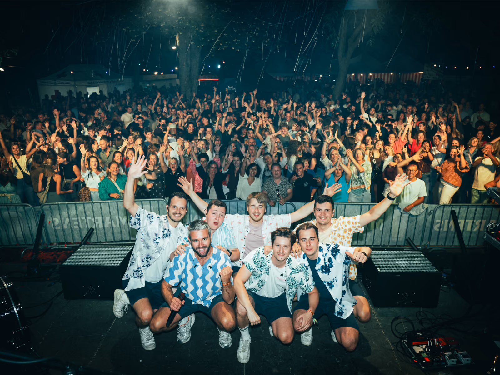 Zeven mannen in zomerse overhemden poseren op een podium voor een groot, juichend publiek tijdens een nachtelijk evenement.