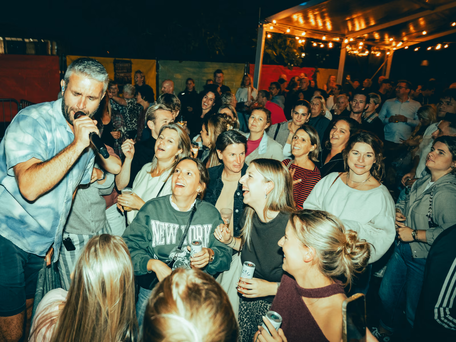 Mensenmassa geniet van live muziek bij een avondfeest buiten onder stringverlichting, terwijl een man met een microfoon dicht bij het publiek optreedt.