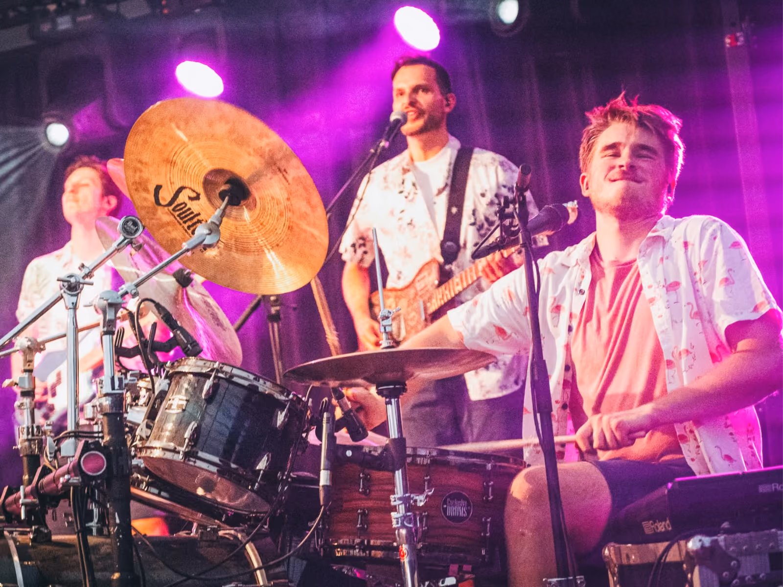 Drummer playing drum set with focused expression while two band members sing and play guitar on stage under purple lighting.