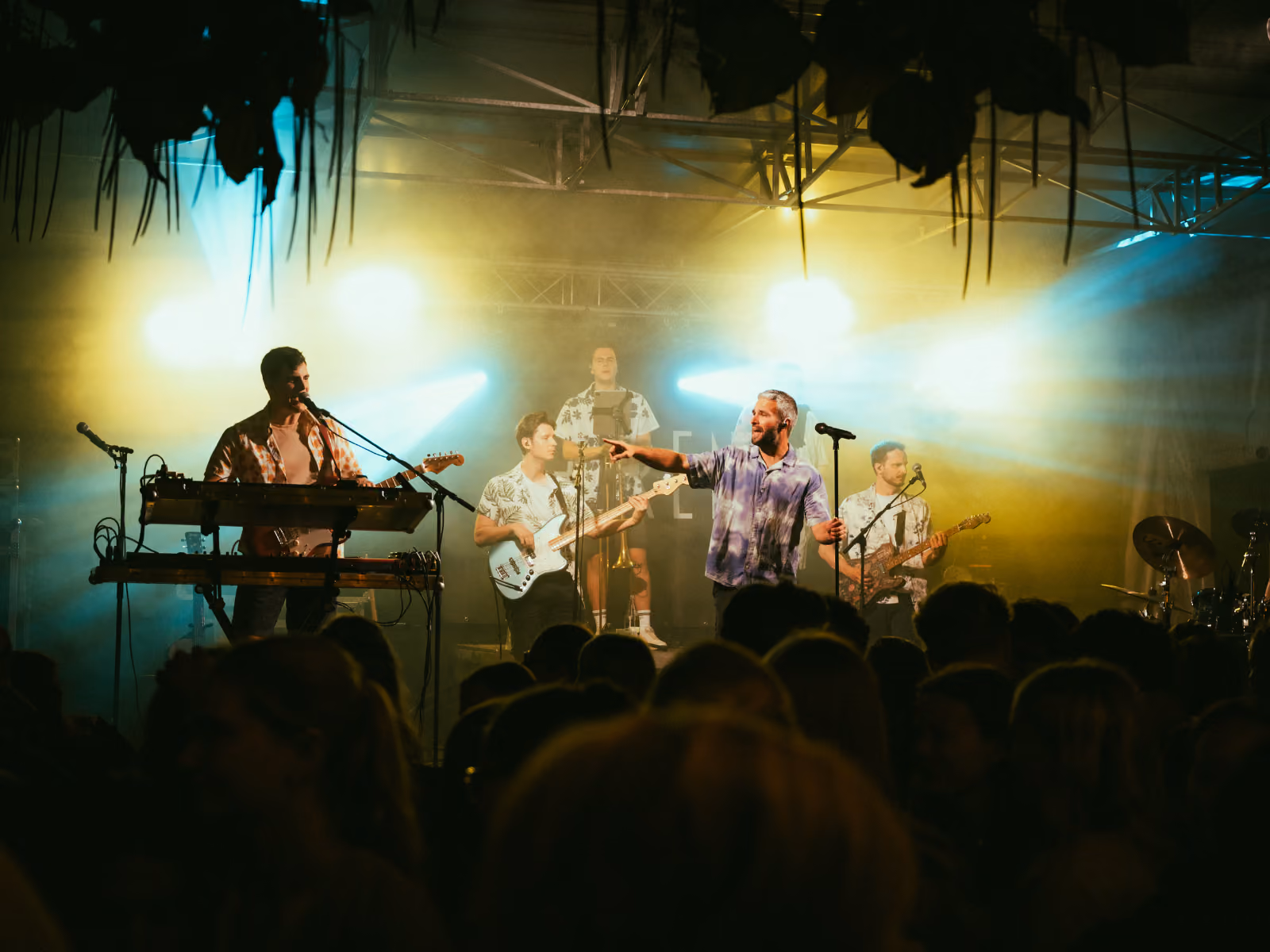Een band speelt live op een podium met felle achterlichten; het publiek kijkt toe terwijl de zanger in het midden met een microfoon staat en in zijn richting wijst.