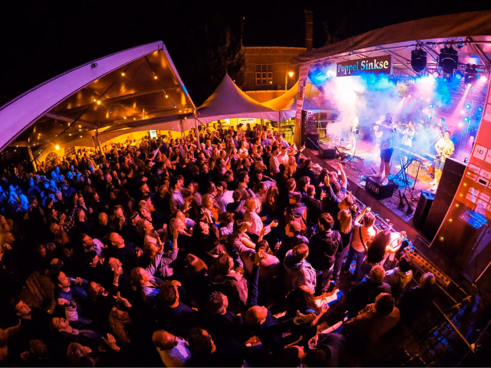 Een groot publiek dat zich verzameld heeft voor een avondconcert buiten onder verlichte partytenten, met een band die op een podium speelt onder gekleurde lichten en rook.