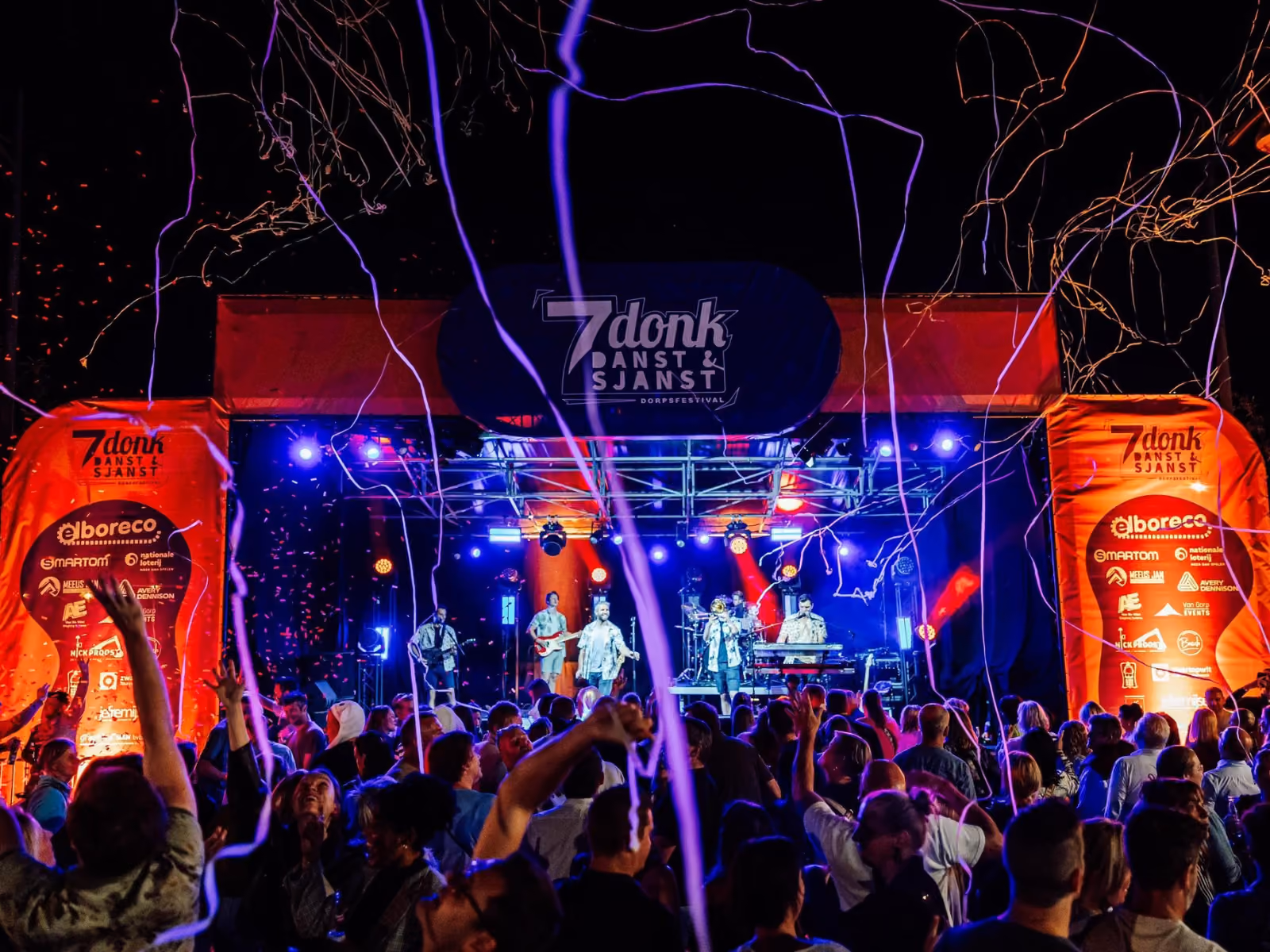 Een nachtelijk muziekfestival met een band die optreedt op een verlicht podium, omringd door een publiek dat geniet en meedoet terwijl kleurrijke lichtstralen en confetti in de lucht hangen.