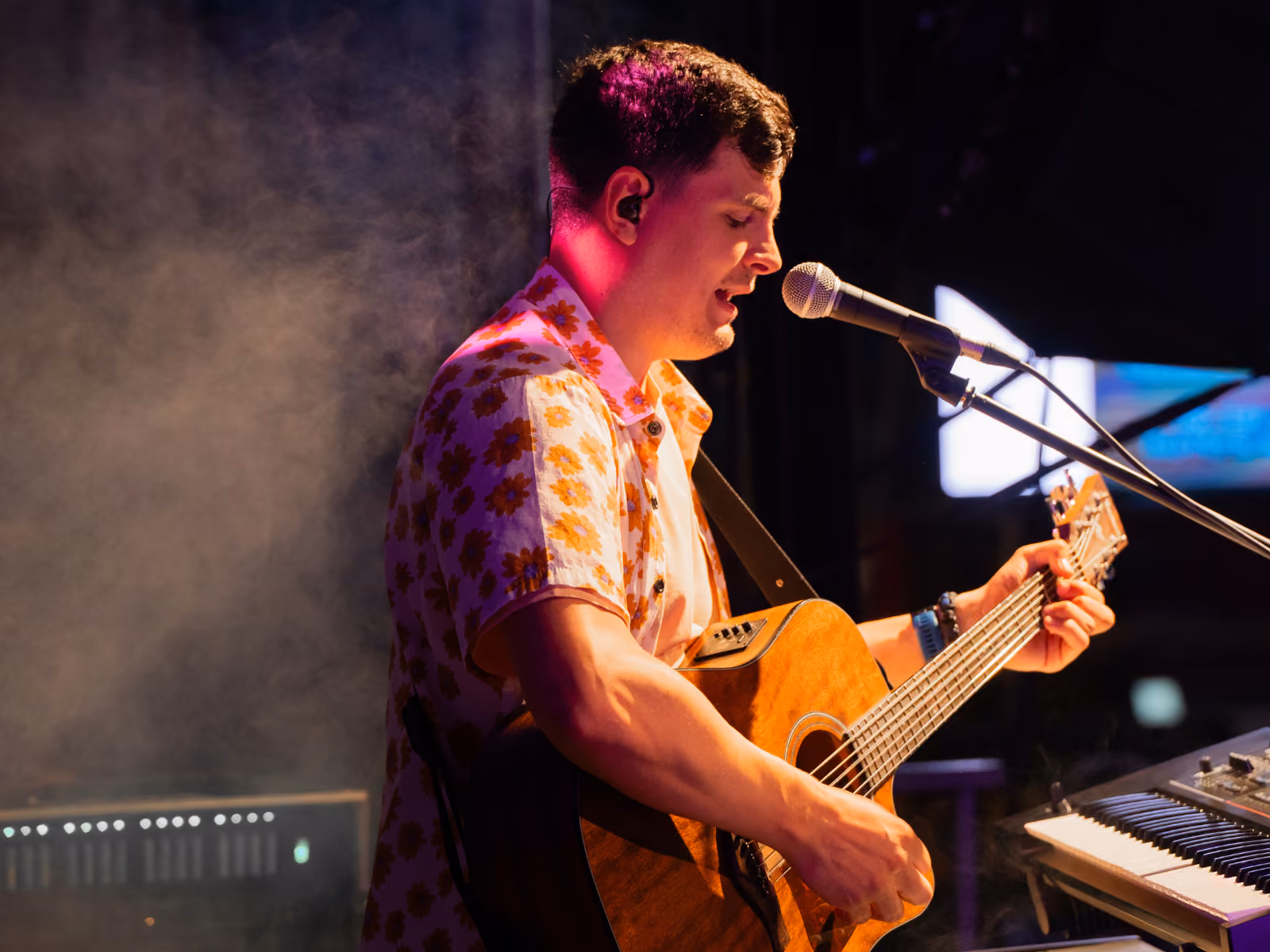 Man zingt en speelt akoestische gitaar op een podium met microfoon en toetsenbord in lage, sfeervolle verlichting.