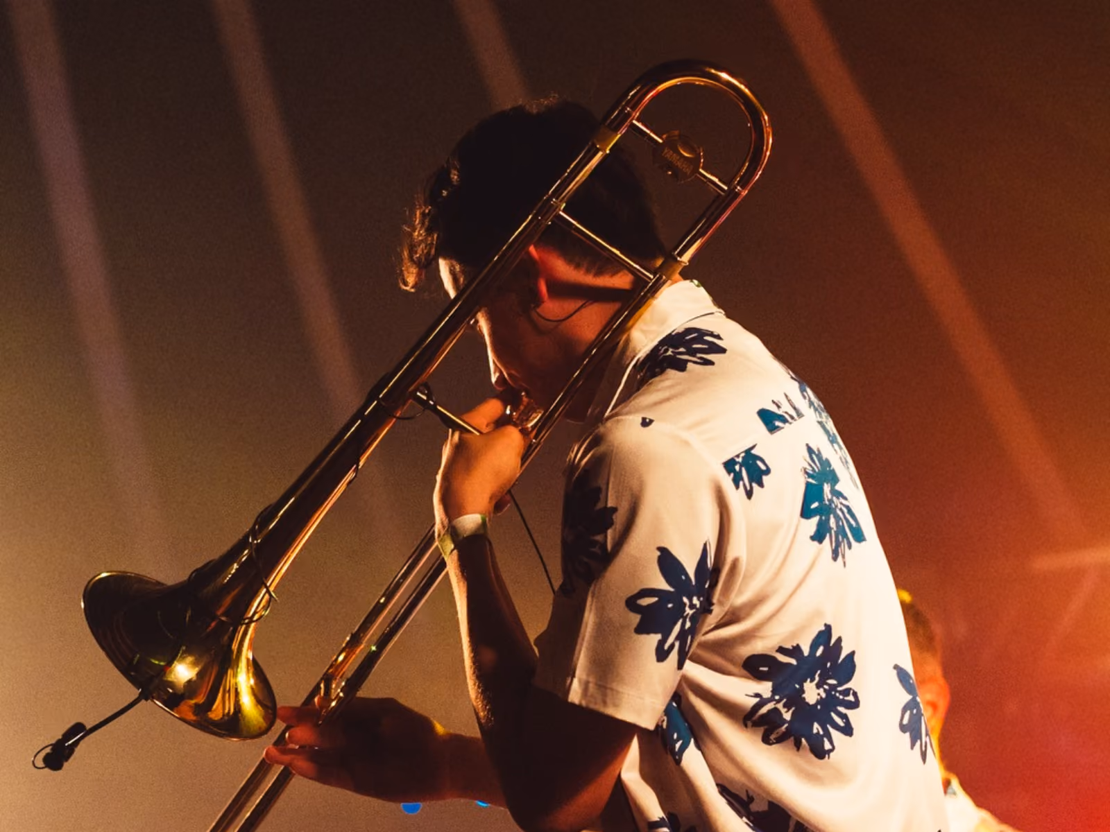 Muzikant speelt trombone op podium, gekleed in een wit shirt met blauwe bloemenprints, verlicht door warme, oranje tinten.