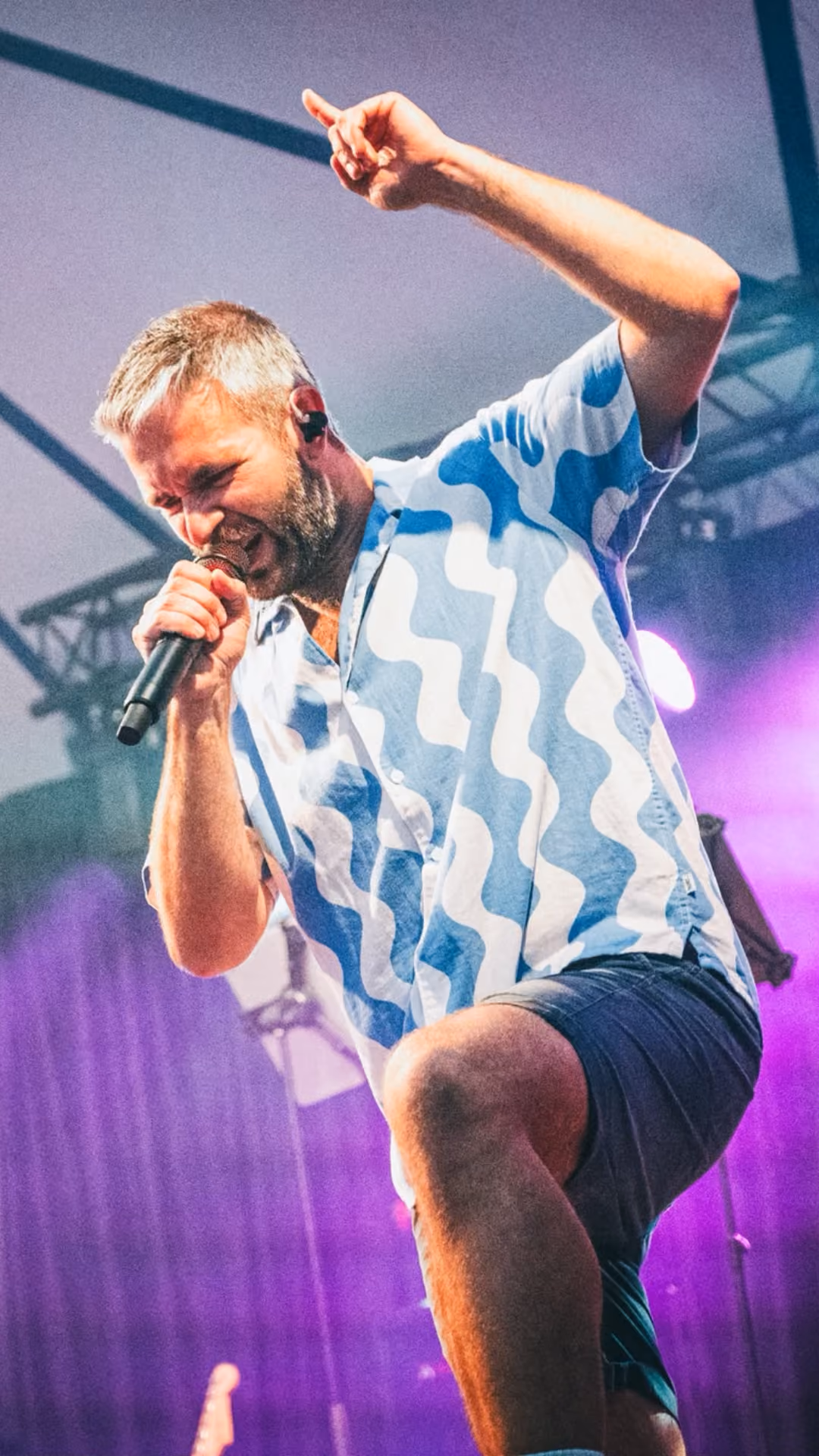 Een man met kort grijs haar en een baard die op het podium vol passie in een microfoon zingt; hij draagt een blauw-wit shirt met een golvend patroon en een donkere korte broek, met één knie omhoog en podiumverlichting op de achtergrond.
