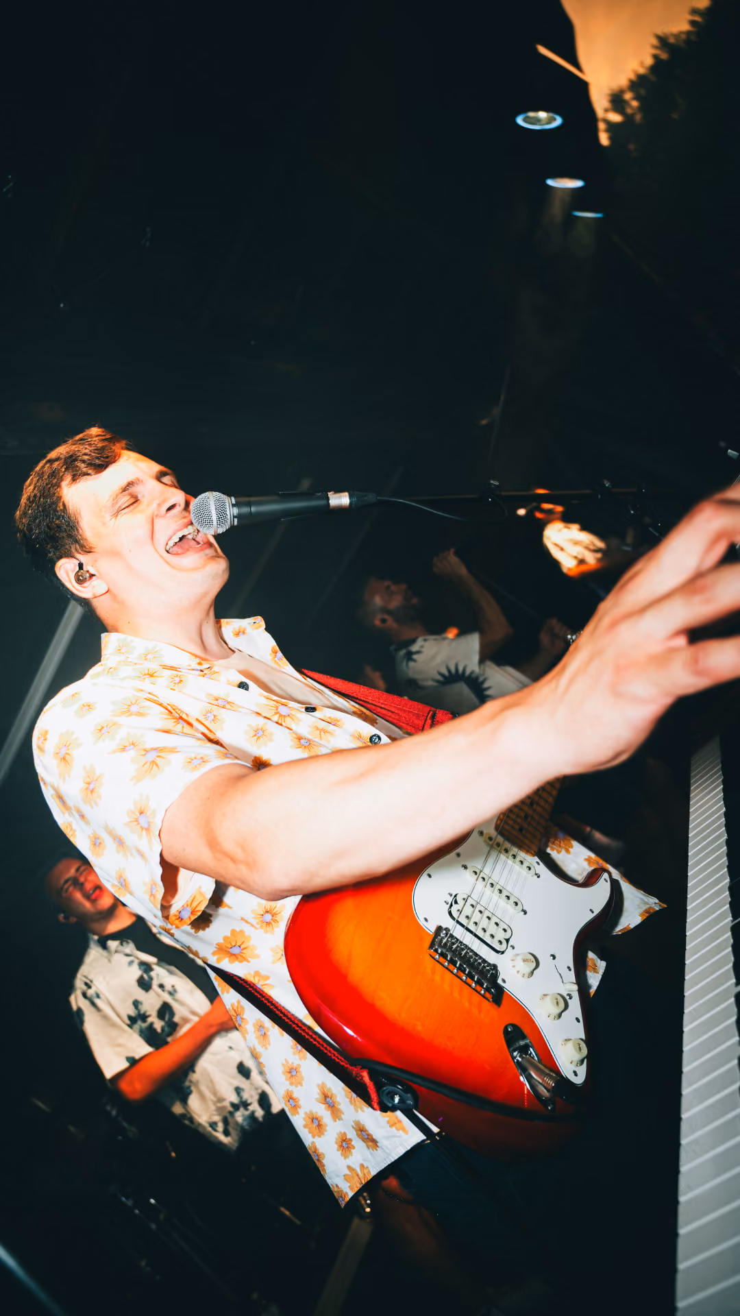 Man zingt gepassioneerd in een microfoon terwijl hij een elektrische gitaar speelt op een podium met twee andere muzikanten op de achtergrond.