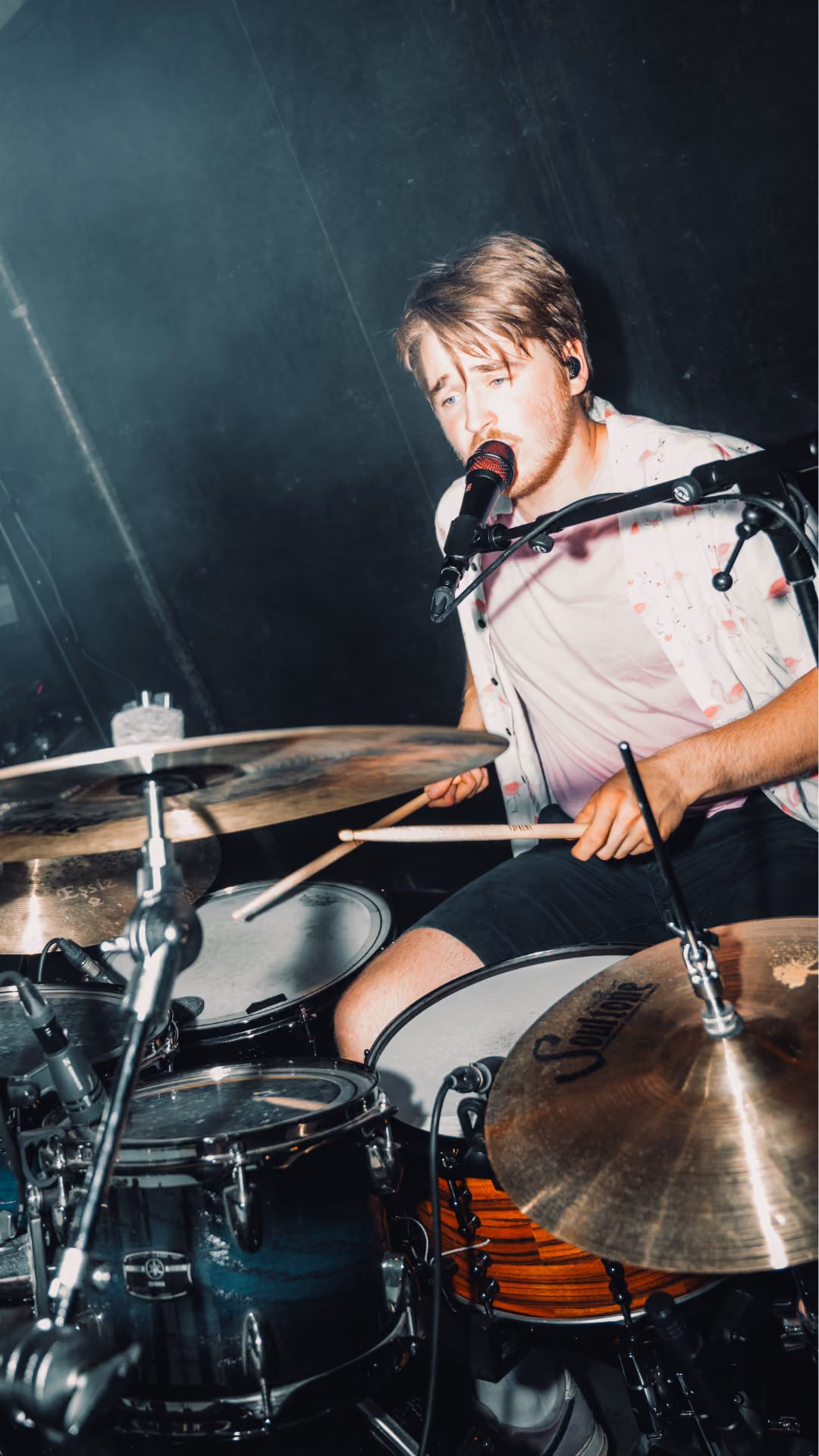 Man speelt drumstel en zingt in een microfoon tijdens een live muziekoptreden, omringd door verschillende drumonderdelen en microfoons.