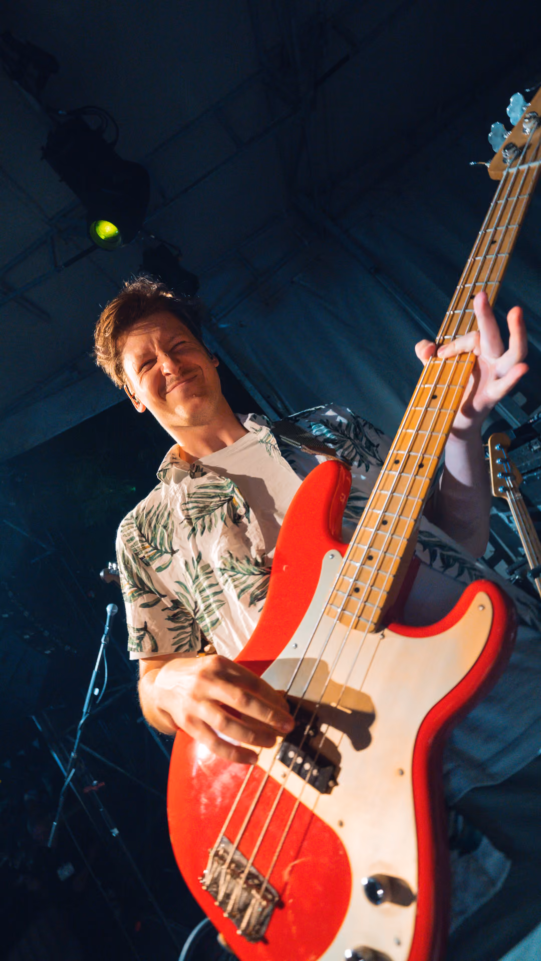 Een man in een lichtgekleurd shirt met groene bladmotieven die op het podium onder blauwe verlichting op een rode elektrische basgitaar speelt.