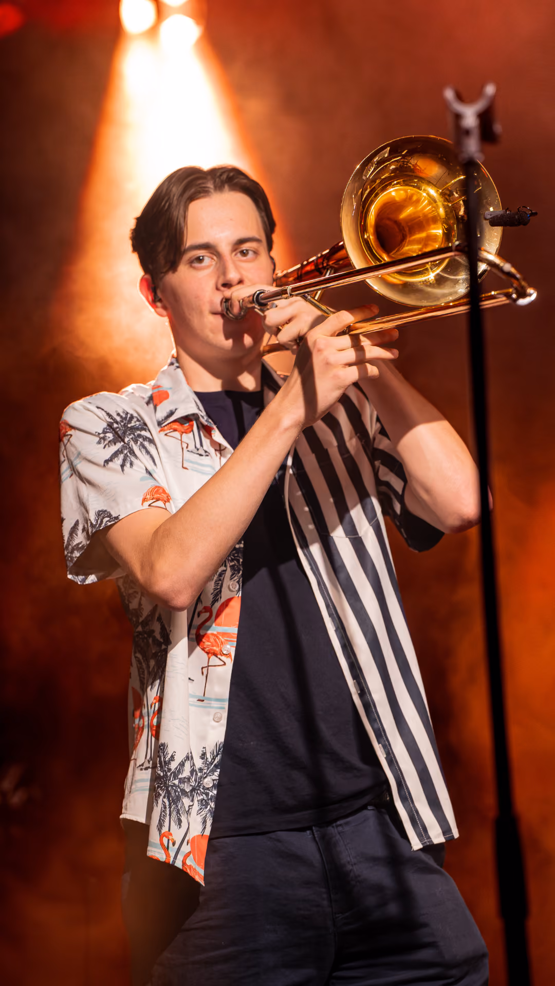Jonge man speelt trombone op een podium met warme oranje en rode verlichting op de achtergrond.
