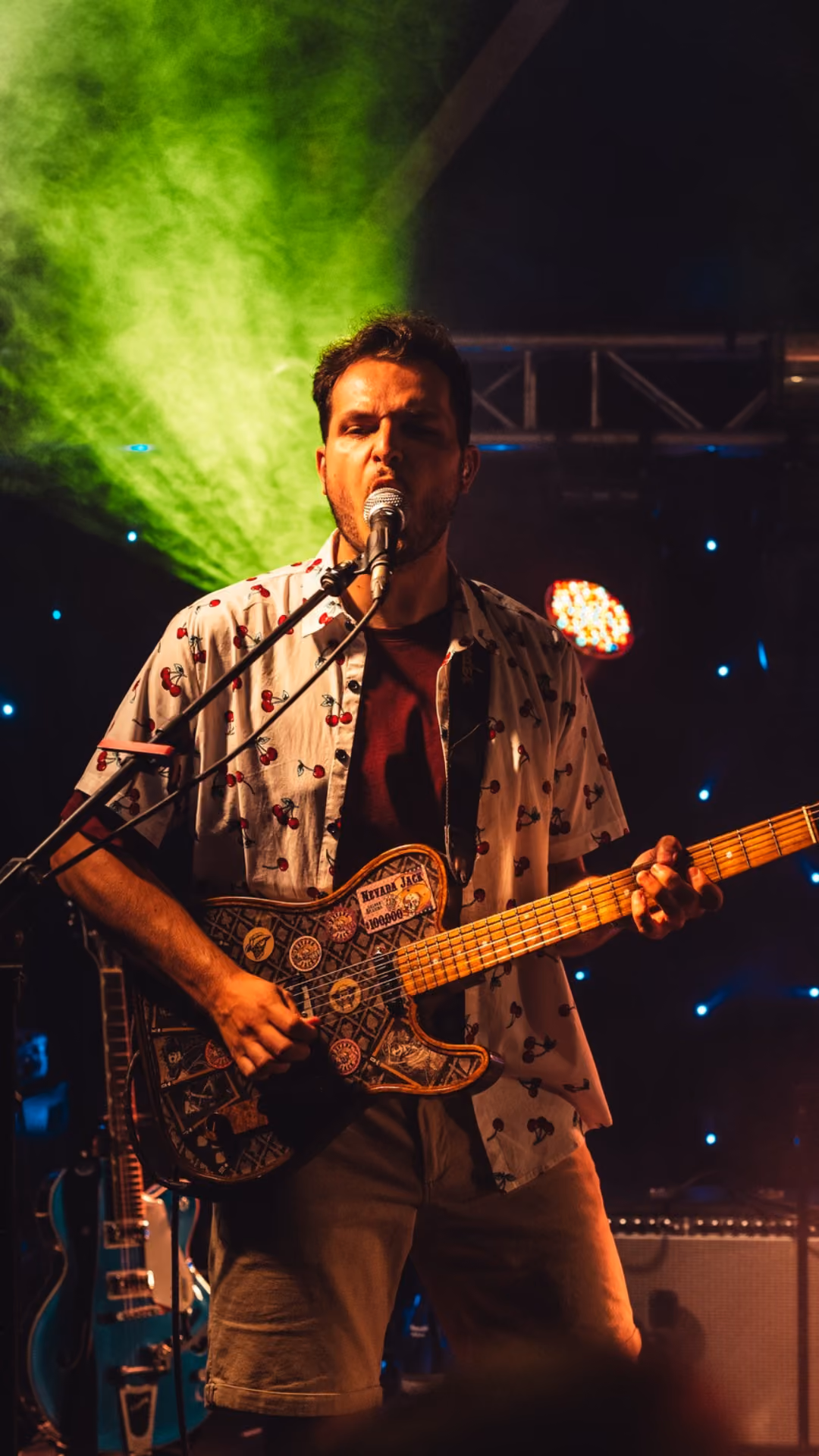 Man zingt en speelt gitaar op een podium met groene lichtstraal en kleurrijke verlichting op de achtergrond.