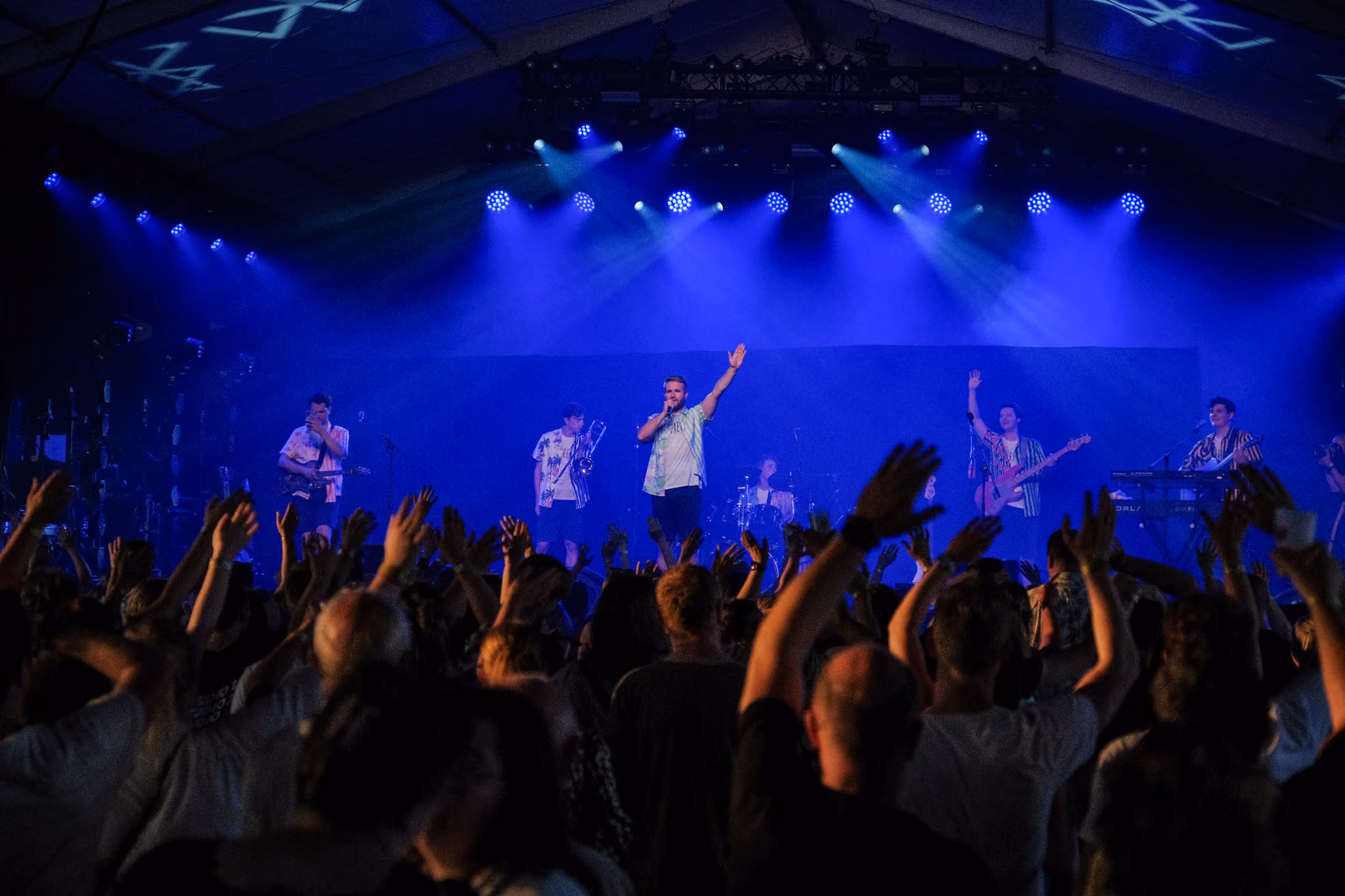 Band speelt op een podium onder blauw licht terwijl het publiek met opgeheven handen meejuicht tijdens een concert in een overdekte ruimte.