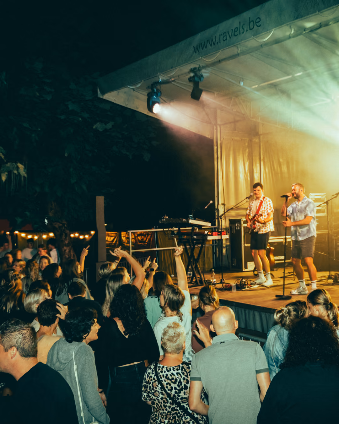 Live muziekoptreden op een verlicht podium met twee muzikanten, een zanger met microfoon en een gitarist, terwijl een enthousiast publiek in de avonduren toekijkt en meedoet.