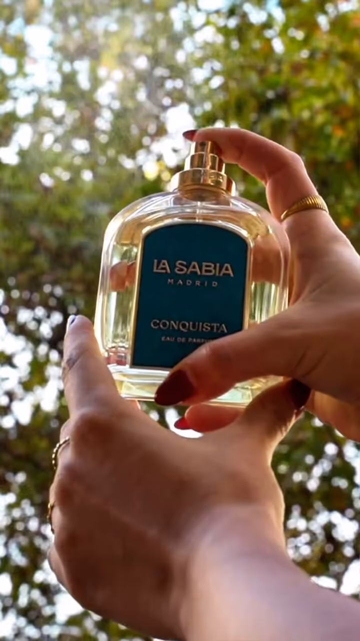 Hands holding a La Sabia Madrid Conquista perfume bottle outdoors against blurred green foliage.