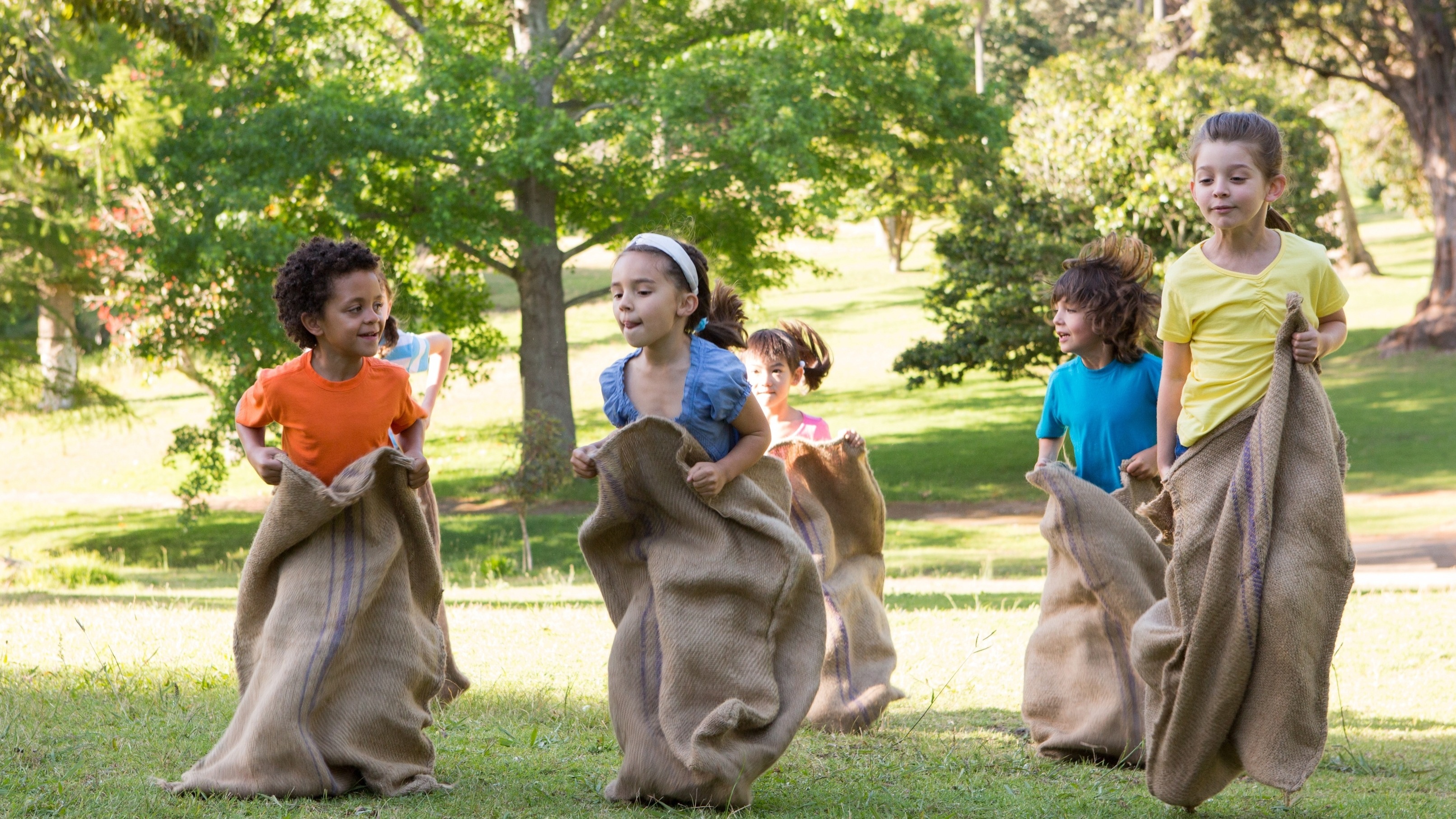 Five children racing in burlap sacks across the grass in a sunny park