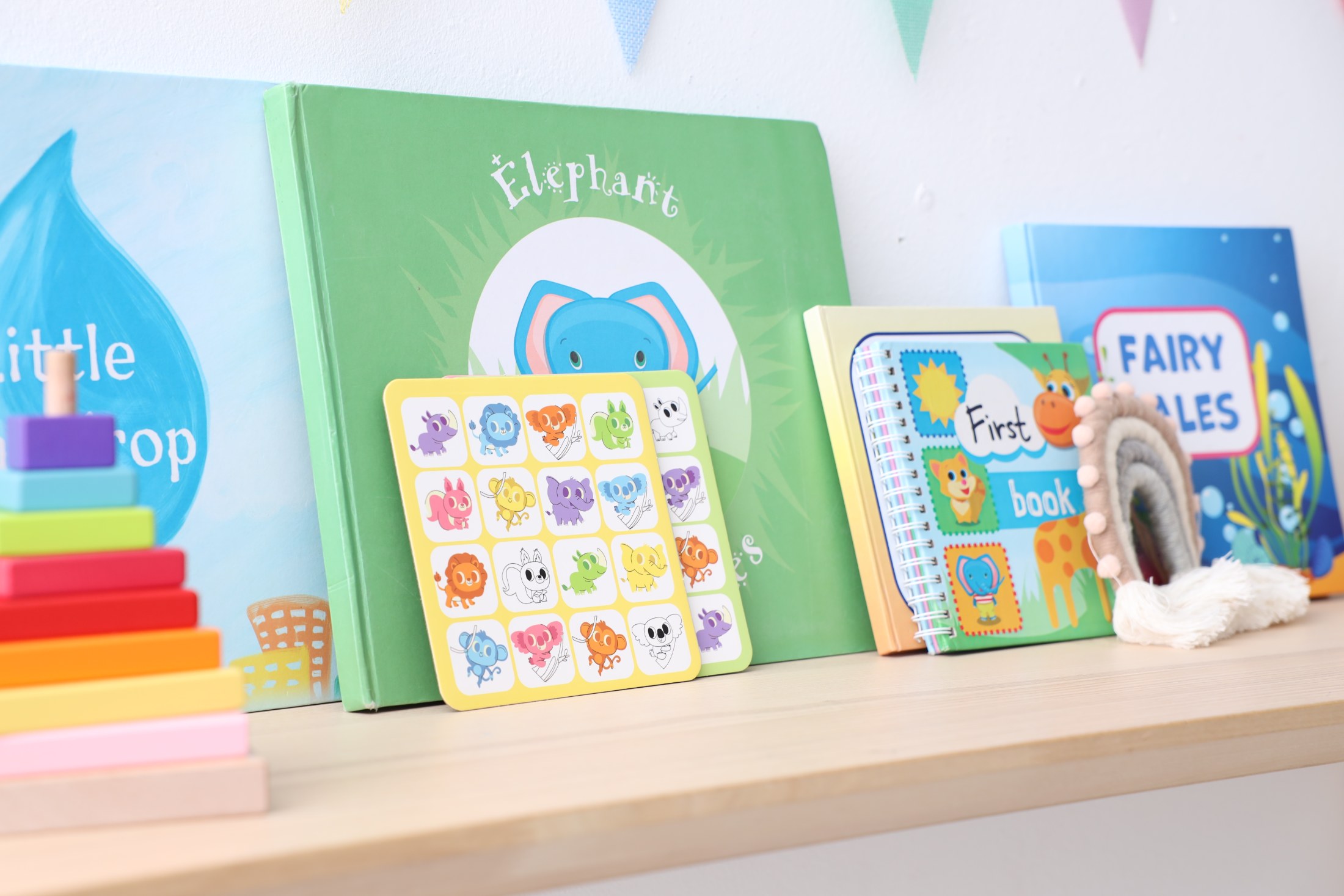 A bright and cheerful close-up shot of a light wooden shelf adorned with various children's books and educational toys. Visible items include a green book featuring an elephant, a blue fairy tales book, and a spiral-bound notebook labeled 'First book'. A colorful rainbow-shaped toy with white tassels and a multi-colored stacking pyramid add a playful touch to the scene. The natural, soft lighting enhances the vibrant colors of the illustrations, creating a warm and inviting atmosphere suitable f