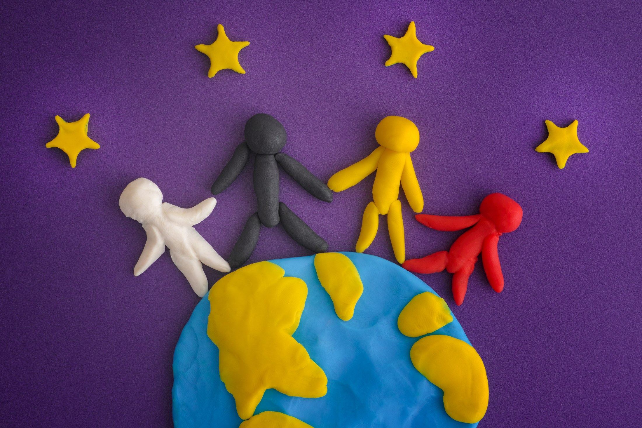 Four colorful clay figures are standing on a clay earth with yellow continents on a blue background with five yellow stars. The figures are white, black, yellow, and red. The figures all have outstretched arms and legs and are posed in the same way.