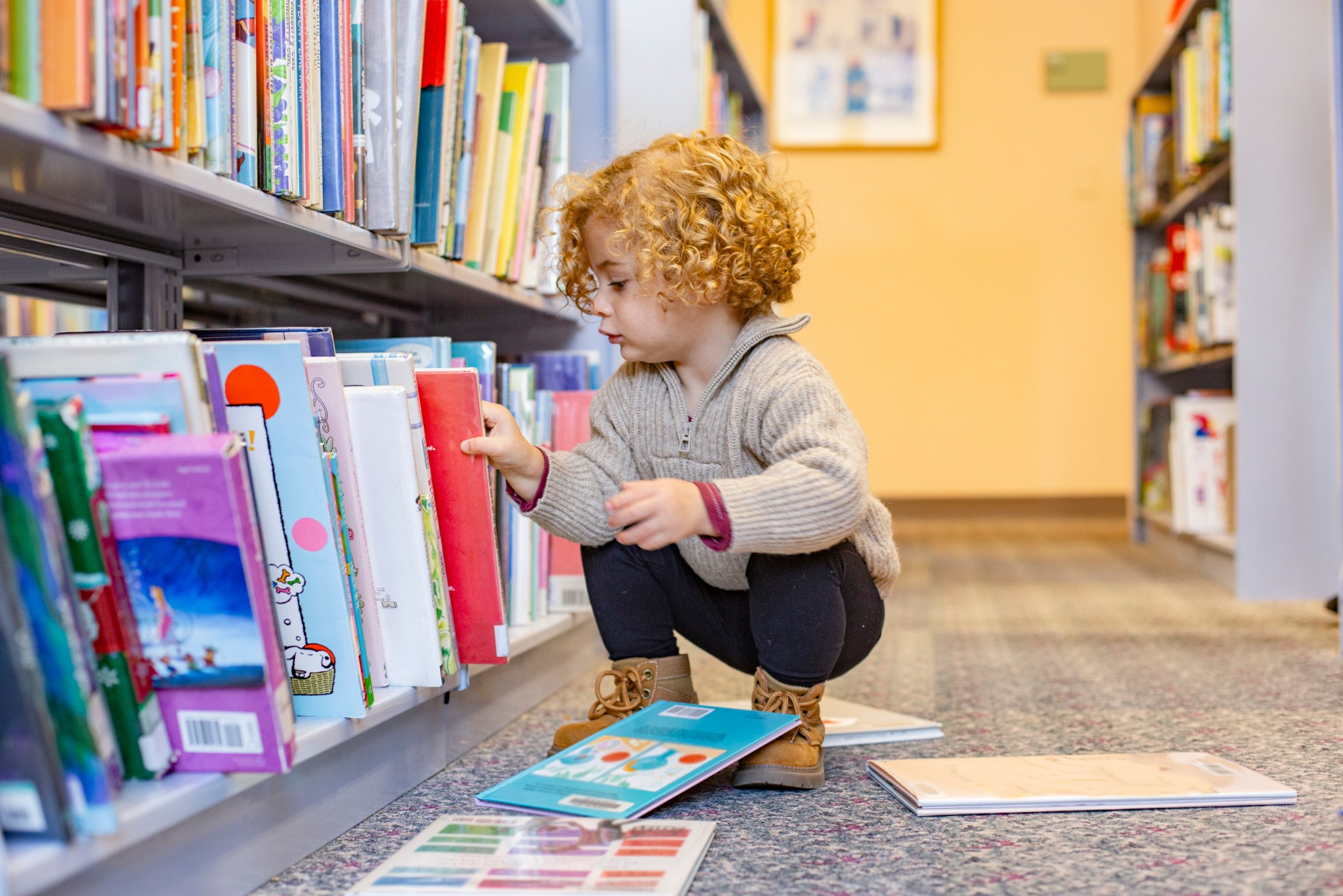 In a brightly lit library, a toddler with curly reddish hair is absorbed in selecting a book from a low shelf. The child wears a beige sweater and dark leggings, squatting to examine the colorful array of books. Several open books lie on the floor, suggesting an active exploration of stories. The setting is a well-stocked library with rows of books extending into the background, emphasizing a quiet and educational environment. The photo's composition and natural lighting evokes a mood of curiosi