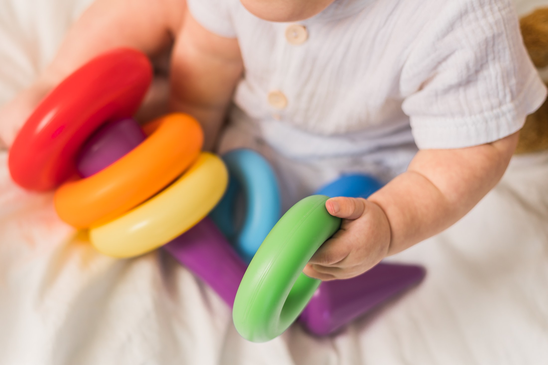 A close-up shot shows an infant playing with a toy made up of colorful, circular rings. The hands of the infant grasp a green ring, part of the larger toy. The other rings are red, orange, yellow, blue, and purple. The baby is wearing a white, buttoned shirt. The scene is set indoors, on a white blanket.