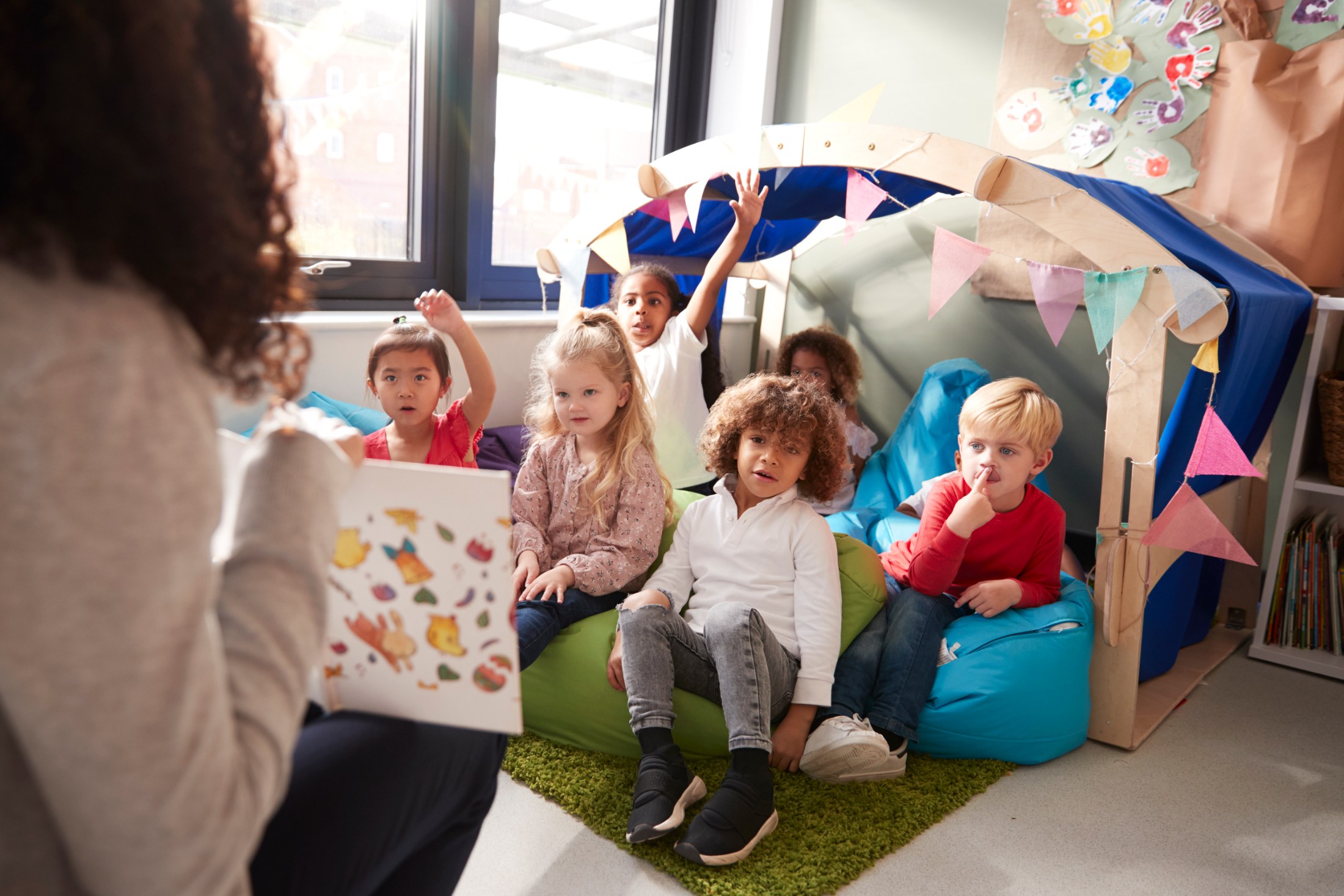 A group of seven children are gathered in a classroom setting, with their hands raised in excitement as they listen to a woman reading a book. The children are seated on colorful beanbag chairs arranged on a green rug. A small decorative tent stands behind them, adorned with pennant flags. The setting is bright, with natural light streaming in through a window. This lighthearted photo is well-suited for use in educational materials, children's books, or marketing campaigns related to early child