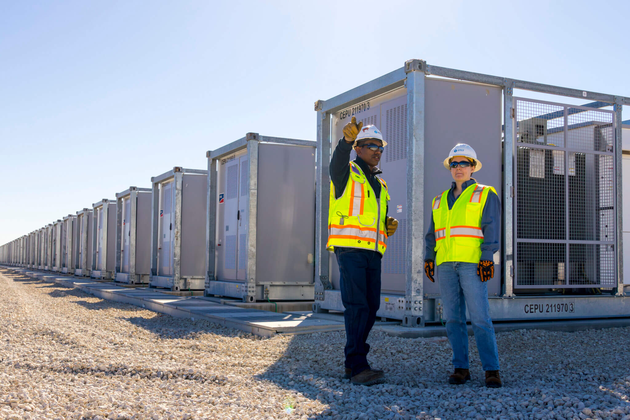 Two workers in safety vests, helmets, and gloves standing on gravel beside large industrial metal containers in a row under a clear blue sky, with one worker pointing ahead.
