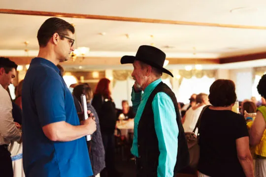 Two men engaged in conversation at a social gathering, one wearing a blue shirt and glasses, the other in a turquoise shirt and black cowboy hat, with other people mingling in the background.