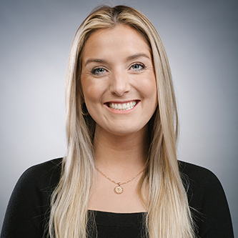 Smiling young woman with long blonde hair wearing a black top and gold necklace against a neutral gray background.