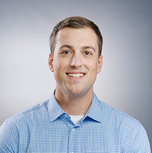 Portrait of a smiling young man with short brown hair wearing a light blue checkered shirt over a white undershirt, against a plain light gray background.