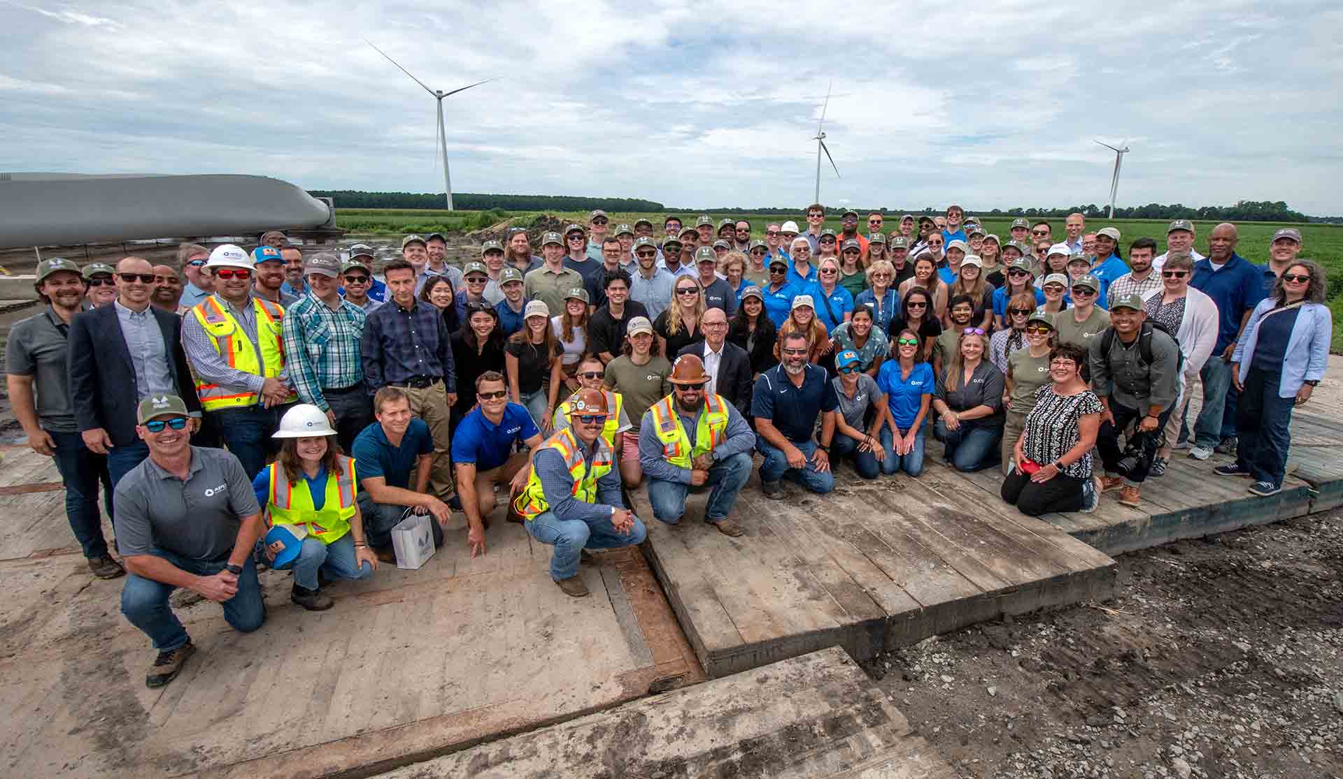 Large group of people posing outdoors on wooden platform near wind turbines under a cloudy sky.