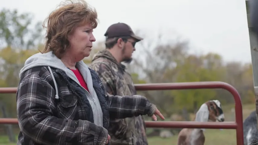 Woman in plaid jacket and man in camo jacket standing near red fence with a goat behind them.