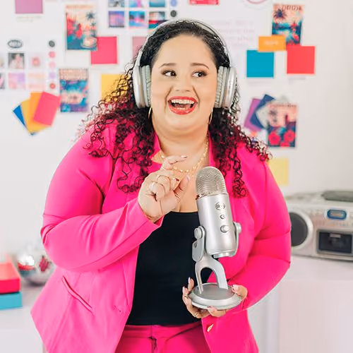 Kristine Gonzalez wears large headphones and smiles while speaking into a silver microphone in a vibrant, design-filled office.