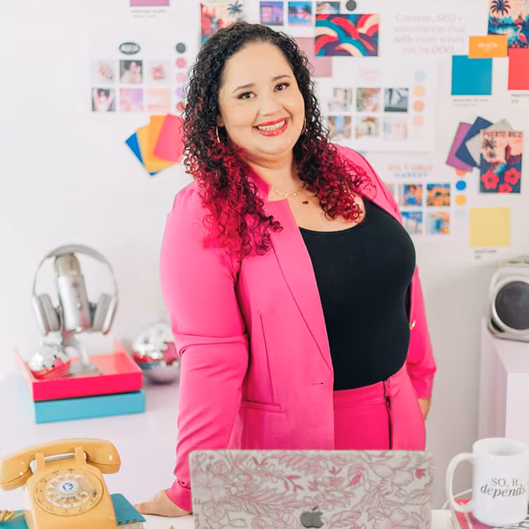 Kristine Gonzalez stands confidently at her desk in a bright, creative workspace, wearing a vibrant pink suit with a black top. Behind her is a colorful mood board filled with branding materials, swatches and inspiration images, highlighting her bold, design-forward aesthetic.