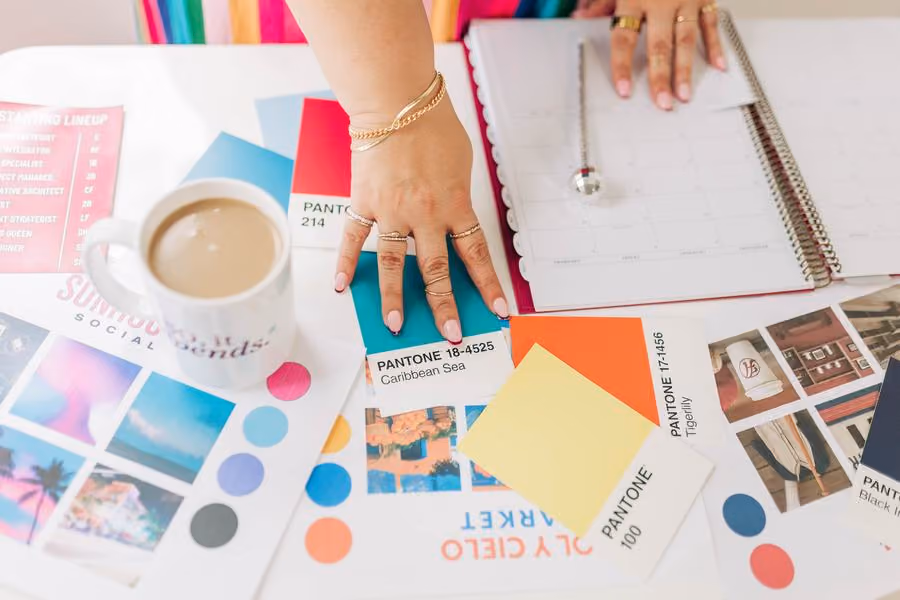 Overhead view of Kristine Gonzalez's hands on a planner beside color chips, printed layouts and a coffee