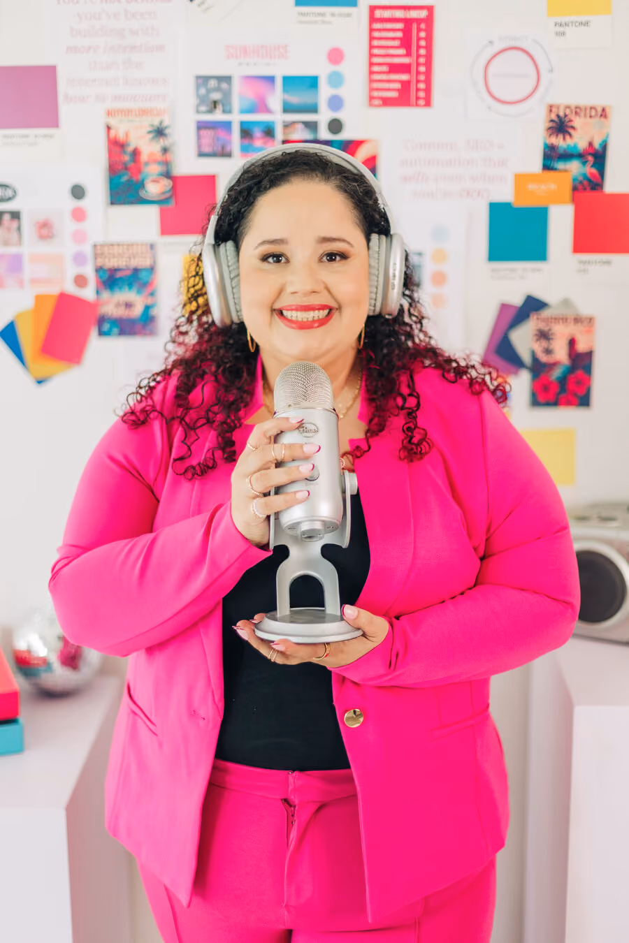 Kristine Gonzalez in a pink suit and headphones holding a podcast microphone