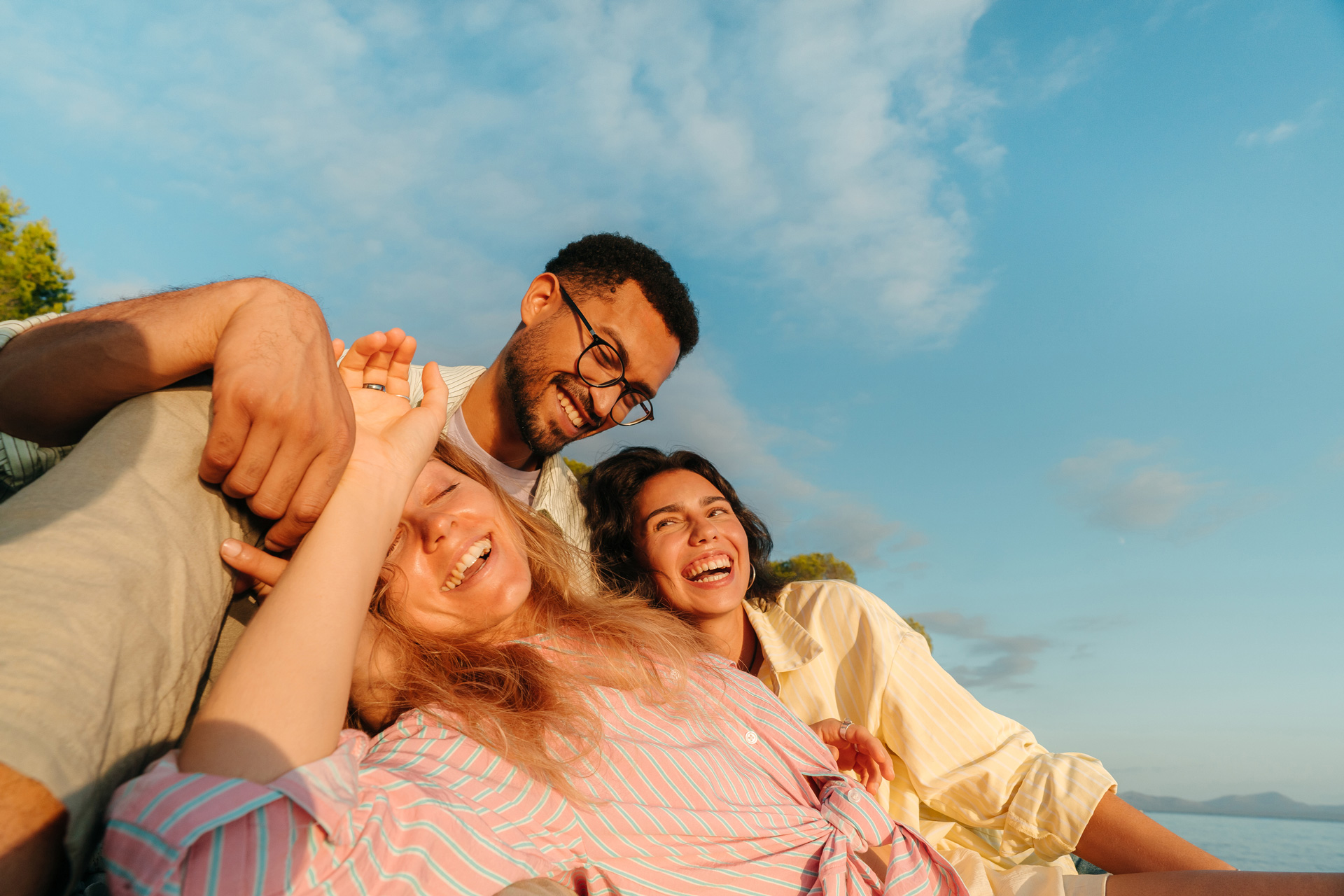 Three friends outdoors laughing and smiling under a blue sky with light clouds, with two women in striped shirts leaning against a man wearing glasses and a light shirt.