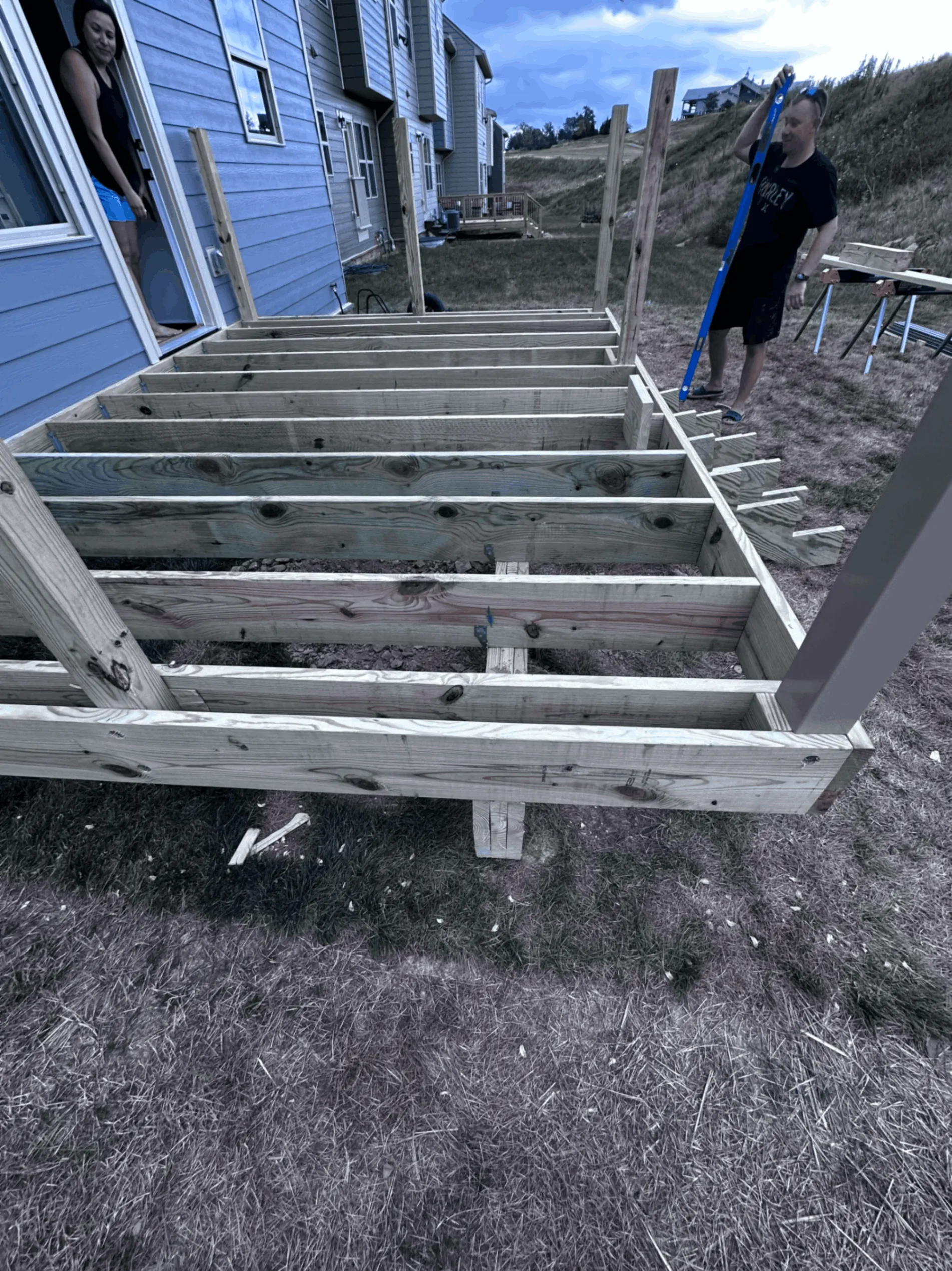 Pressure-treated deck framing under construction behind a blue home