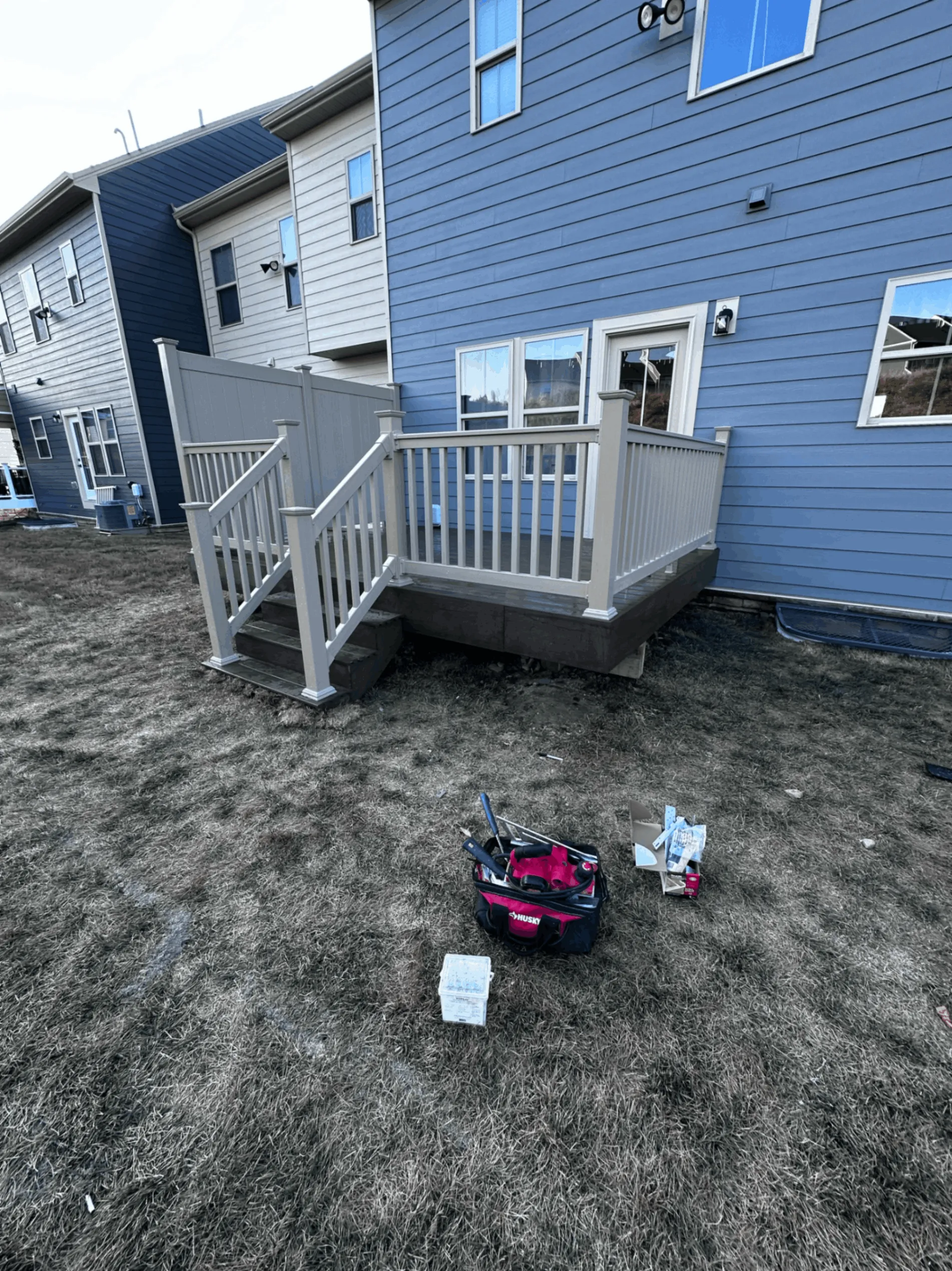 Small gray deck with white railings behind a blue sided townhome
